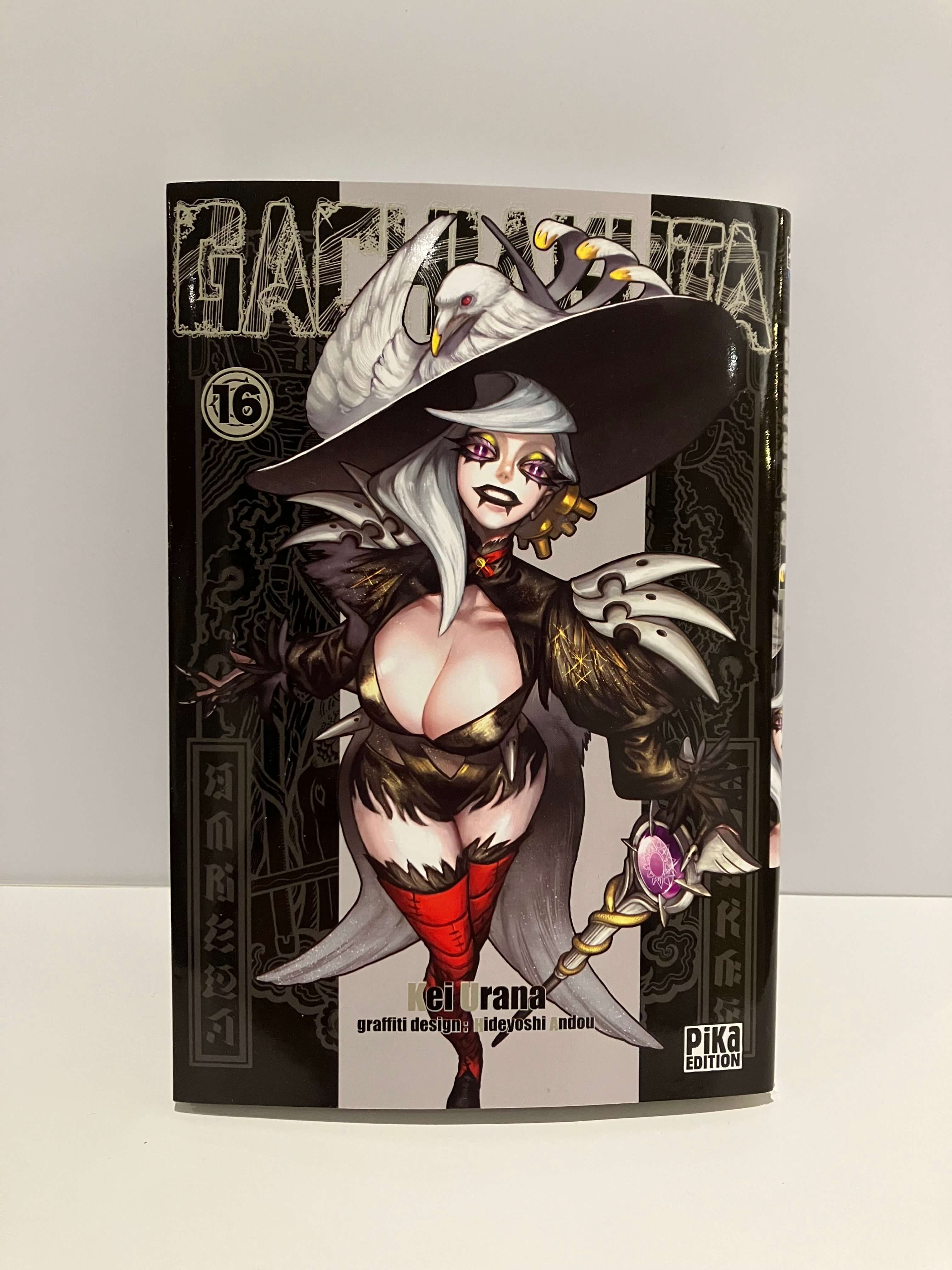 Couverture de manga Gachiakuta tome 16 : une femme aux cheveux blancs en tenue sombre et chapeau pointu tient un sceptre, sur fond noir aux motifs détaillés.