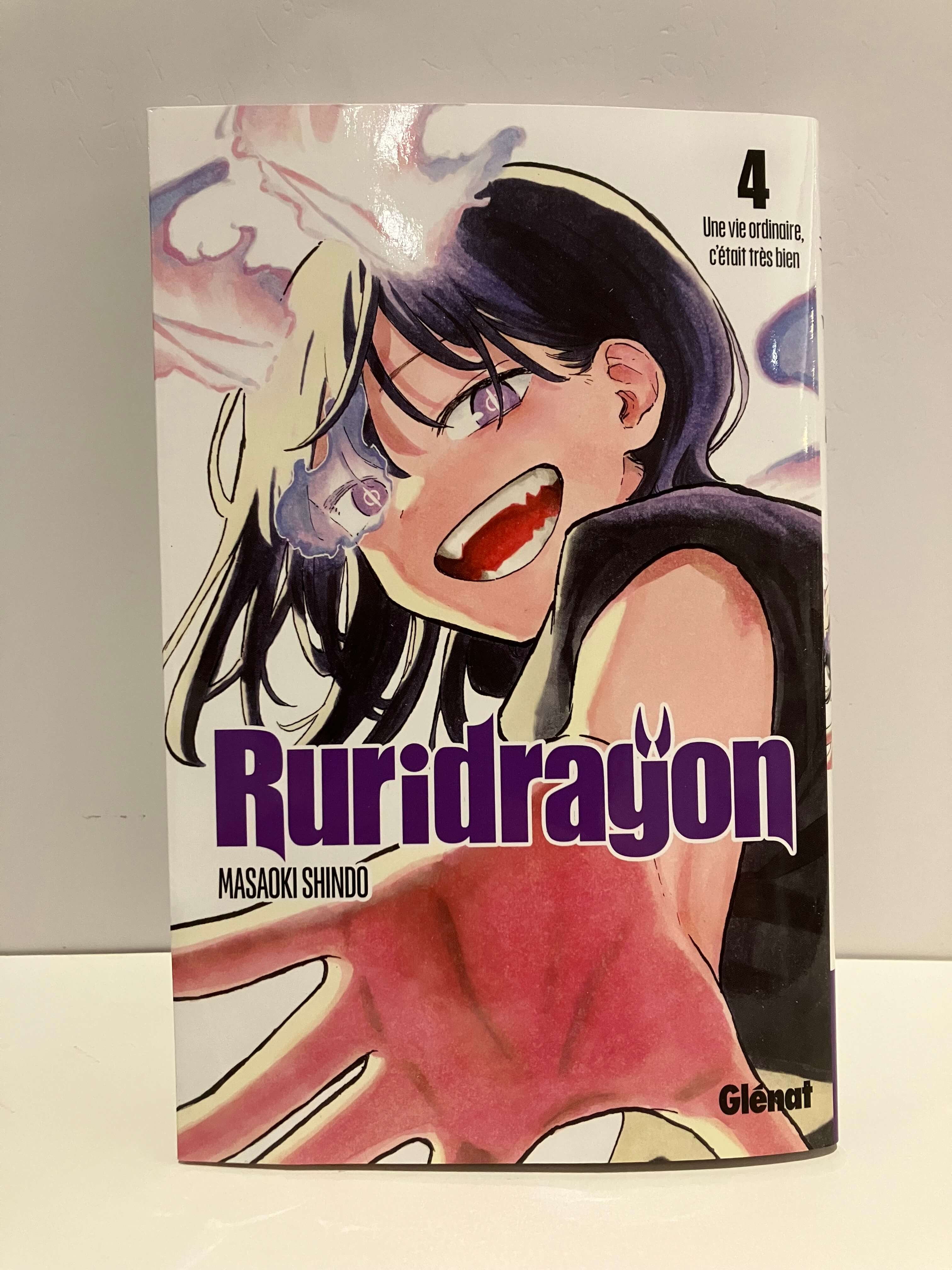Couverture de manga Ruridragon tome 4 : Une jeune fille aux cheveux noirs avec une mèche claire occupe presque toute la couverture, affichant un large sourire et tendant la main vers le lecteur. Des volutes violettes s’échappent de son visage, accentuant l’aspect fantastique de l’illustration. Le titre Ruridragon apparaît en grandes lettres violettes au centre, sur un fond blanc aux touches pastel.
