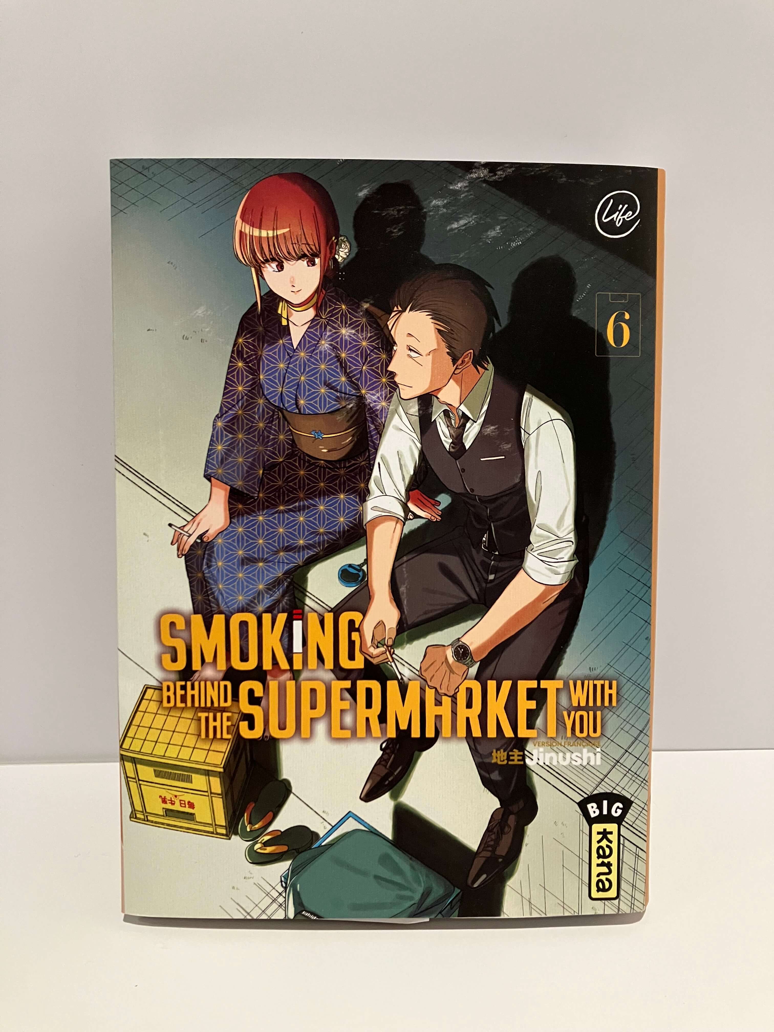 Couverture de manga Smoking Behind the Supermarket with you Tome 6 : Une jeune femme en kimono bleu à motifs et un homme en chemise blanche avec gilet noir sont assis côte à côte près d’une caisse en plastique jaune, dans une ruelle sombre derrière un supermarché. L’ambiance nocturne et intimiste est renforcée par les ombres projetées sur le mur derrière eux.